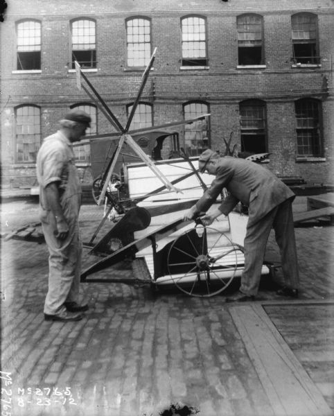 Two Men Discussing Binder Two men are standing in the factory yard discussing a binder. The man on the left is wearing overalls and a cap, and the man on the right is wearing a cap and suit. There is horse-drawn wagon and a brick factory building in the background.