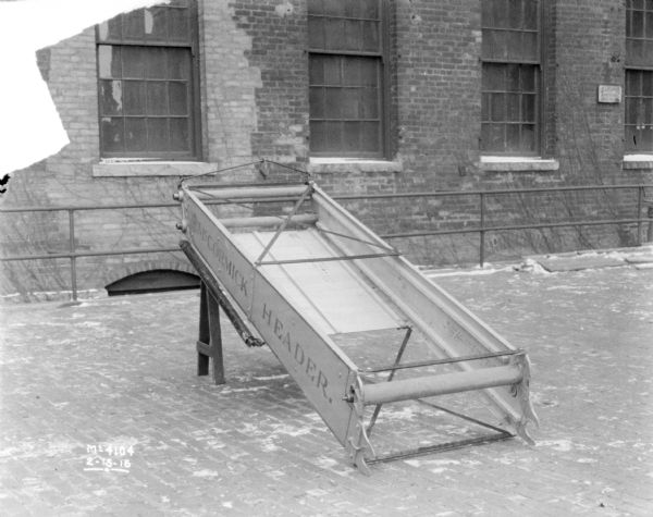 McCormick Header Box set-up on a sawhorse outdoors in the factory yard. There is a railing and a brick factory building in the background.