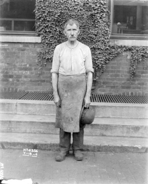Man Wearing Work Apron Full-length portrait of a man standing in front of a brick building. He is wearing a work apron and is holding a hat in his left hand.