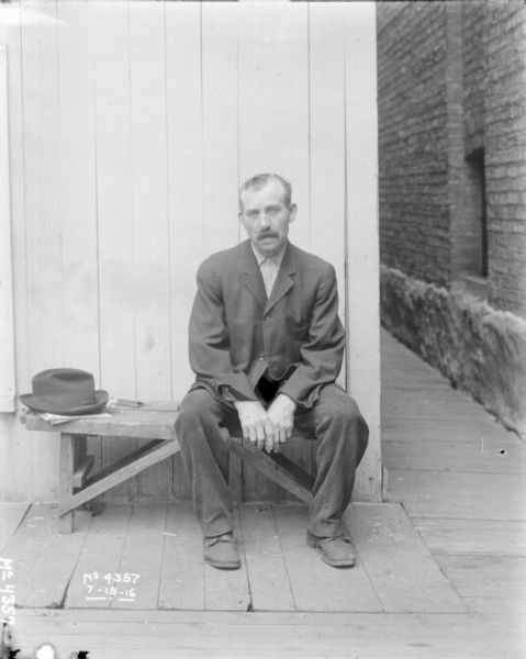 Man Wearing Suit Sitting on Bench Man wearing a suit sitting on a bench outdoors, with his hat on the bench nearby. The side of a brick building is on the right.
