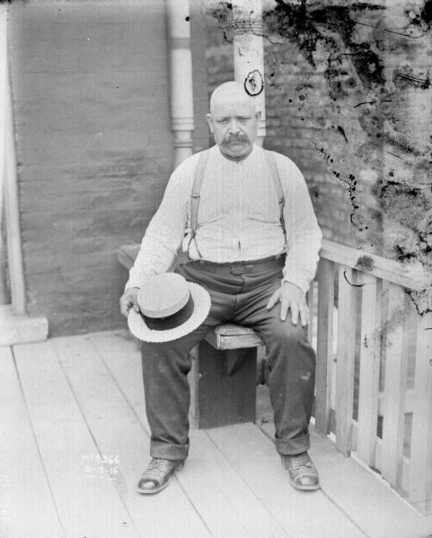 Man Sitting on Porch Full-length portrait of man wearing a shirt, suspenders and pants sitting on a bench on a porch. He has a straw hat on his lap.