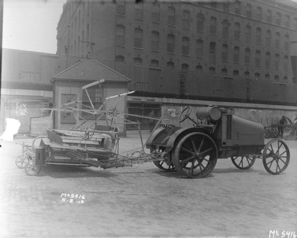 Tractor-Drawn Binder at McCormick Works Tractor-Drawn Binder outdoors in factory yard. An elevated railroad platform and brick factory buildings are in the background. Men are working in the background on the right.