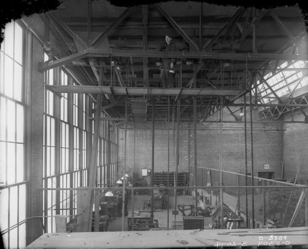 Man on Catwalk Above Manufacturing Area View looking up towards a man standing on a catwalk high above the manufacturing area. He is holding an oil can, and may be working on maintaining the belt-driven machinery attached to the ceiling of the factory. A tall length of windows is along the left wall, and skylights are on the right. Men are working below on the factory floor.