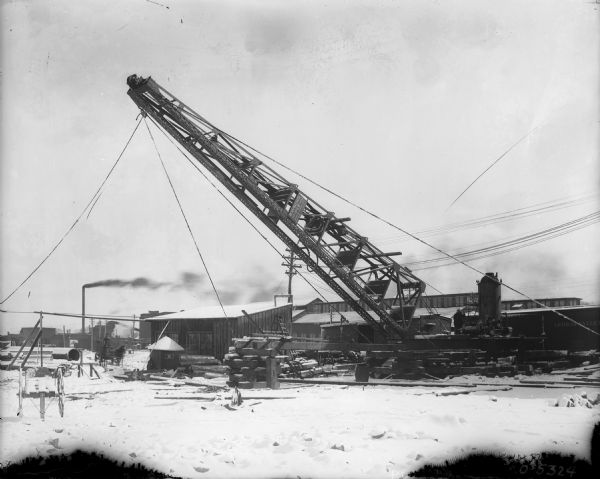 Large Crane at Factory View of factory yard with a large crane near buildings and railroad cars. One of the railroad cars has a sign that reads: "Lehigh Valley." The sign on the crane reads: "Raymond Pile Concrete Company, Engineers and Contractors, New York, Chicago." There is factory building in the background, and a smokestack is streaming dark-colored smoke. Men are working near the base of the crane on the right, and other men are on a horse-drawn wagon on the left. Snow is on the ground.
