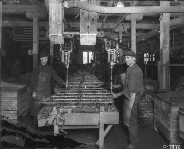 Men Assembling Products on Assembly Line Men are assembling products, Spring tooth harrows, on an assembly line. An American Flag is hanging from the ceiling in the background on the left.