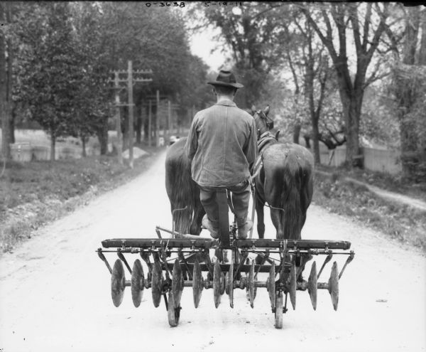 Horse-Drawn Disk Harrow on Transport Truck Rear view of a man transporting a Disk Harrow with a team of horses down a road.