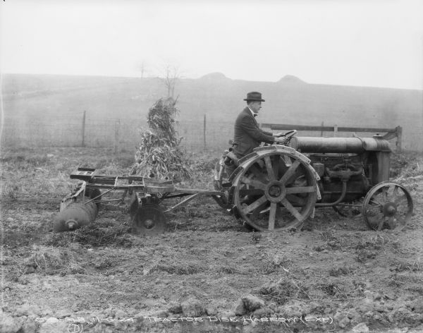 Man Driving Tractor Disk Harrow Right side view of a man wearing a suit and hat driving an experimental tractor Disk Harrow. A cornstalk is behind the Disk Harrow.