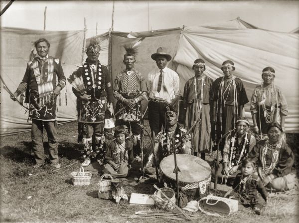 Ho-Chunk performers gathered behind a drum and Winnebago baskets at the 1908 Homecoming. Standing from the left are Jim Swallow (MaPaZoeRayKeKah), William Massey (ChawRoCooChayKah), George Eagle (WaNaKeeScotchKah), Benjamin Thundercloud (NySaGaShiskKah), and Flora Thundercloud Funmaker Bearheart (WaNekChaWinKah). The woman standing on the far right is Susie Lena Pettibone Johnson (ChoNukKaWinKah), and the woman seated to the left is Addie Littlesoldier Lewis Thunder (KzunchJeKahRayWinKah). Thomas Thunder (HoonkHaGaKah) is seated beside her behind the drum. Remaining names are unknown.