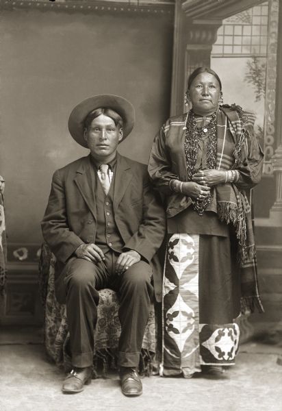 Studio portrait of George Monegar (EwaOnaGinKah) and Fannie Stacy Lincoln Monegar (LeBaWinKah) posing standing in front of a painted backdrop. He is wearing a suit jacket, vest, tie, and hat, and she is wearing beads, a long embroidered skirt, and wool shawl. Fannie Stacy Lincoln Monegar is the mother of Frank Lincoln according to the notes of Jackson County Historical Society, Wisconsin.