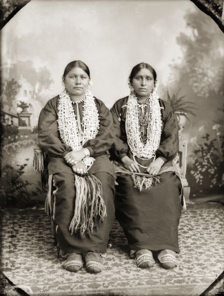 Full-length studio portrait in front of a painted backdrop of two unidentified women wearing silver earrings, shell necklaces, finger-woven wool sashes, and moccasins with beaded top flaps.