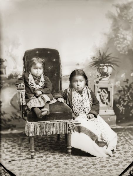 Studio portrait of two Ho-Chunk girls posing in front of a painted backdrop. A small Ho-Chunk girl is posing sitting in a tasseled arm chair on the left, and is wearing several bugle beads. The other Ho-Chunk girl is posing sitting, probably a low stool on the right, and is wearing several bugle beads and earrings, with a Hudson's Bay trade blanket on her lap.