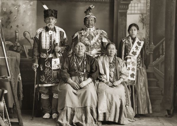 Studio portrait of two Ho-Chunk women sitting in front of two Ho-Chunk men and a Ho-Chunk woman posing standing, all wearing regalia. The group is in front of a painted backdrop. The women sitting and standing are wearing several necklaces and earrings. The standing woman and men also wear ribbon work clothing, and the man standing on the left is holding a medicine bag. Identified standing are John Hazen Hill (HaNaKah), left, Alec (Alex) Lonetree (NaENeeKeeKah), and Mary Clara Blackhawk, daughter of Lucy Emerson Brown; sitting are Lucy Emerson Brown, left, and Lucy Long-Wolf Winneshiek (ShunkChunkAWinKah).