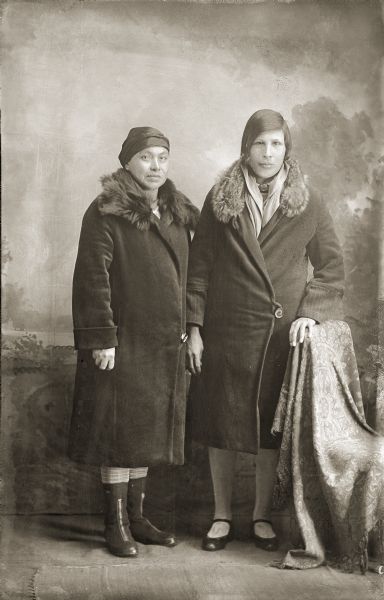 Studio Portrait of Dora Thunderking Lowe and Jennie Youngthunder Decorah Full-length studio portrait in front of a painted backdrop of two Ho-Chunk women posing standing. They are wearing winter coats with fur collars. Identified as Dora Thunderking Lowe (AkSeKahHoNoKah), right, and Jennie Youngthunder Decorah (PetHakChaCooWinnKah).