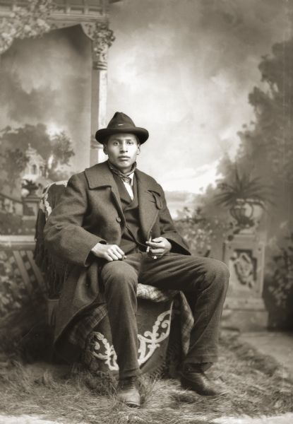 Full-length studio portrait of Will Thunder (NaKikSayWaHeKah) (son of [WaConChaKah] John Thunder aka Dr. Thunder and [WeHonPeKaw] Lucy Bear, Thunder) sitting on an embroidered blanket, draped on a chair, that appears in many of Van Schaick’s photographs. He is wearing a suit, tie, and overcoat, and is holding a cigarette. This blanket was not Ho-Chunk, but a studio prop. In the background is a painted backdrop.