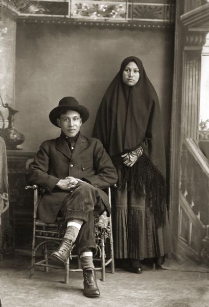Studio Portrait of John Falcon and Lucy White/Waukon Studio portrait in front of a painted backdrop of a Ho-Chunk man posing sitting on the left with crossed legs, and wearing a suit and hat. Behind him on the right is a Ho-Chunk woman posing standing and wrapped in a Comanche fringed shawl, only showing her face. John Falcon (NaHeKah) is seated next to Lucy White/Waukon, the daughter of Ellen Bigbear.