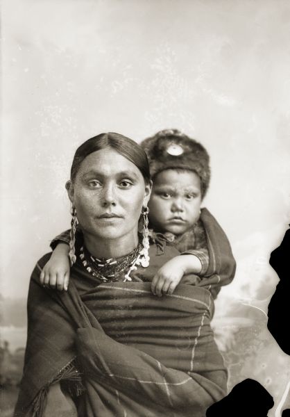 Quarter-length studio portrait of a woman in Ho-Chunk regalia wrapped in a blanket, with a child on her back. The child is wearing a fur hat, and they are posing sitting in front of a painted backdrop. Clara Kingsley (Big) Blackhawk (KeesKawWinKah) carries her son Andrew John (Big) Blackhawk (WaConChaHoNoKah) on her back. Using a shawl as a back sling was a common way to carry infants in this period.