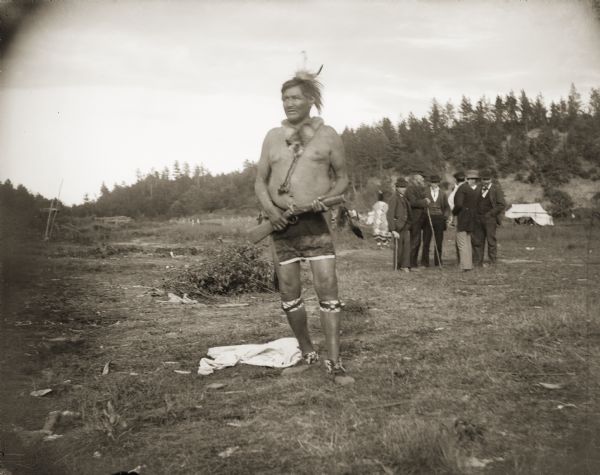 Ho-Chunk man posing standing in a open field holding a flintlock gun. He is wearing garters, moccasins, a breech cloth, necklace, and feathers in his hair. In the background is a group of European American men standing together, and in the far background is a tree-covered hill. He is identified as John Greencloud “Green Thunder” (WauKonChawChoKah), at Frog Place in the Morrison Creek Bottoms at Brown Eagle place. John served in the Civil War as a scout for the U.S. Army with the Comanches.