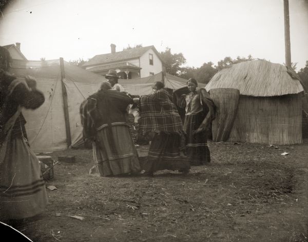 A group of Ho-Chunk women wrapped in shawls gathering at the temporary Ho-Chunk village. Identified as the Homecoming Powwow at the intersection of Main and Second Streets. A house can be seen on a hill in the background.
