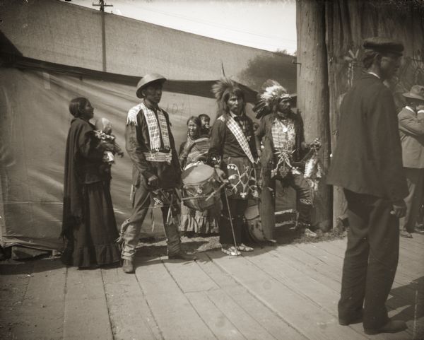 A group of performers taking a break from entertaining tourists. The man on the left is wearing a traditional Ho-Chunk beaded shirt, belt, and breechcloth (apron), as well as a Chinese coolie hat. Coolie hats were used by area farmers and available at the general store downtown. The man in the middle is wearing a porcupine roach headdress with an eagle feather, a Ho-Chunk bandolier bag and beaded apron, and Plains-style moccasins. Identified as probably the Homecoming Powwow at the intersection of Main and Second Streets.
