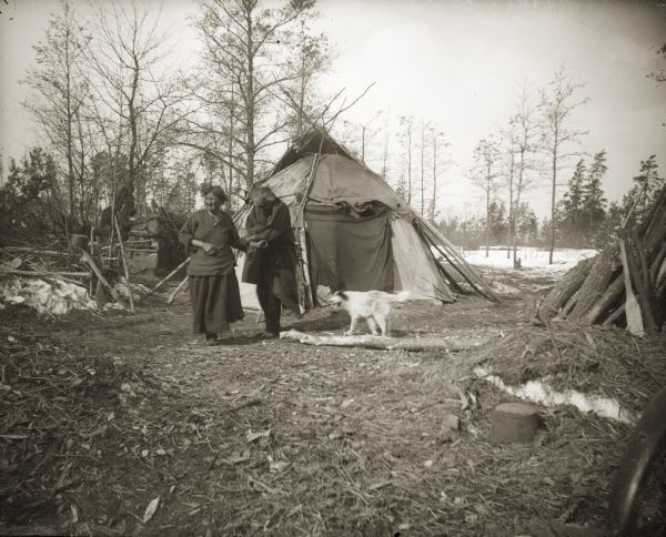 A Ho-Chunk couple and their dog outside of their winter home (ciiporoke). A Hudson Bay blanket is hanging over the door opening. The long tree branches leaning against the canvas were used to open and close the smoke flaps on the top of the roof. On the right foreground is a pile of wood that has been gathered for the winter. The man is reportedly blind.