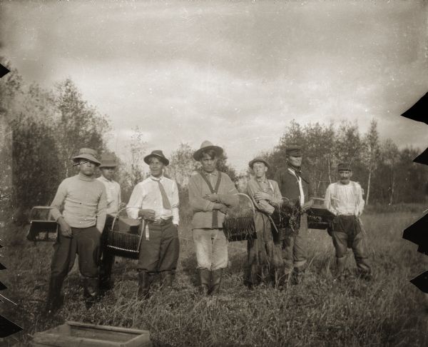 Outdoor group portrait of seven Ho-Chunk men standing outdoors in a cranberry bog, They are holding the hand scoops used to harvest cranberries. The long narrow slats at the front of the hand scoops were used to strip the berries off of the vines. Standing third from left is George Garvin Sr. (WoShipKah); second from right is Jim Swallow (MaPaZoeRayKeKah).