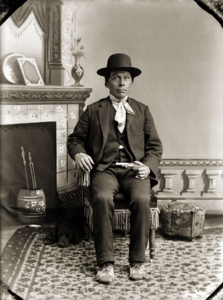 Green Grass (HaWinChoKah), in period dress with the exception of his Ho-Chunk moccasins. He is holding a pistol, and his dog is lying next to his chair on the left.