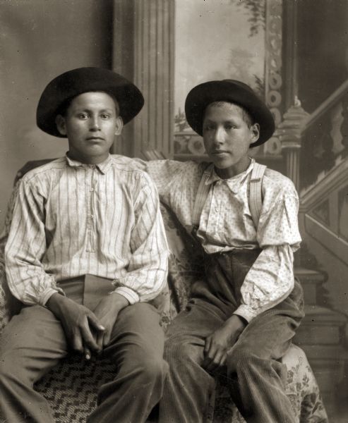 Studio portrait of two Ho-Chunk boys posing sitting in front of a painted backdrop wearing light-colored print shirts and dark hats. The boy on the right has his right arm around the shoulders of the other boy and is wearing suspenders. They are identified as Lou Waukon sitting on the left, and his brother Steve Waukon sitting on the right.