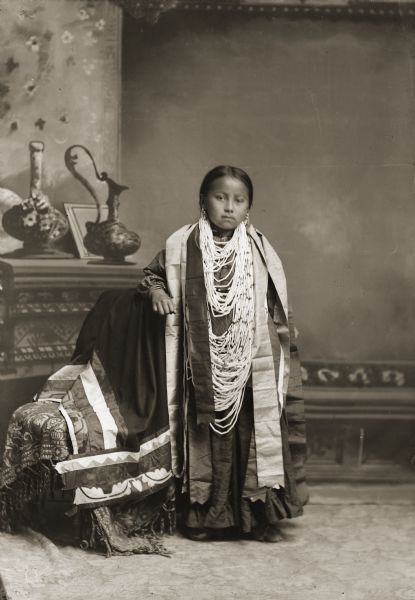 Full-length studio portrait in front of a painted backdrop of a Ho-Chunk girl posing and standing with her arm resting on a chair with a floral applique blanket draped over it. She is wearing a large wampum necklace. 