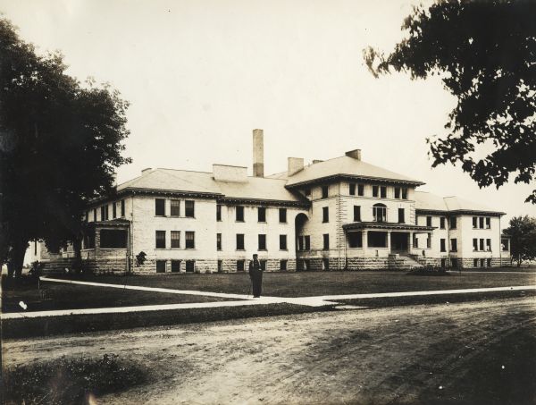 View from road of a building located on the grounds of the Winnebago County Asylum. Standing on a sidewalk in front is a man leaning on a cane.