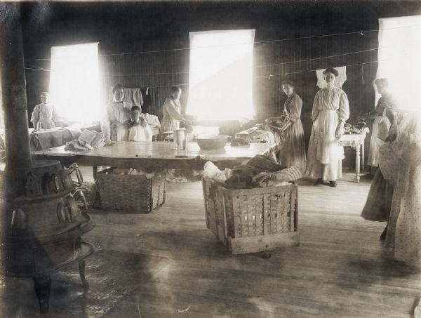 Interior view of the laundry room at the Winnebago County Asylum. Pictured are the staff, including a young boy. Some of the women are ironing articles of clothing. Throughout the room are buckets of laundry, a cast iron stove heating additional irons, and a large table.