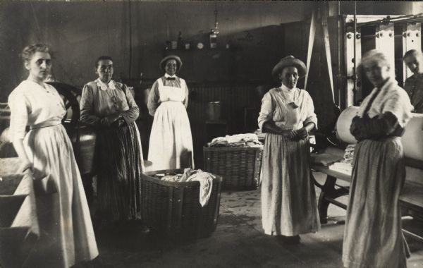 Six staff members pose in the laundry room in the Winnebago County Asylum. In the room are carts of laundry and a machine on the right.