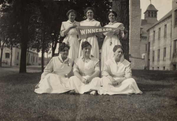 Outdoor group portrait of six nurses, taken on the lawn of the Winnebago County Asylum. Three nurses standing in the back row are holding a "Winnebago" pennant. In the background are large brick buildings.
