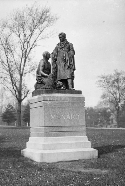 Close-up of a statue honoring Colonel Pierre Menard who also served as the Lieutenant Governor of Illinois and fur traded with Native Americans. Dedicated in 1888, John H. Mahoney's bronze sculpture depicts Menard in a full-length coat and blanket standing over a Native American man who is kneeling, bare-chested, over some pelts. The statue stands in a grassy park.