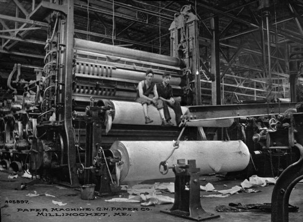 Two men sit on a roller near a large paper machine at Great Northern Paper Company, which was incorporated in 1898 and soon became one of the largest American suppliers of newsprint, mainly on the East Coast. Caption reads: "Paper Machine, G.N. Paper Co. Millinocket, ME."