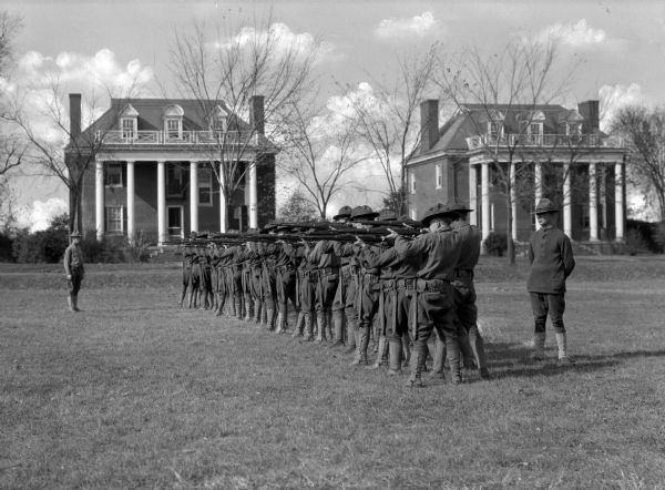 A view of a line of soldiers in the drill field, holding firearms. One officer stands in front of them, and another behind. Two large buildings in the background were built as the officers' quarters of the Washington Barracks in 1901. The Barracks became the Army's center for the training of senior officers to lead large numbers of troops.