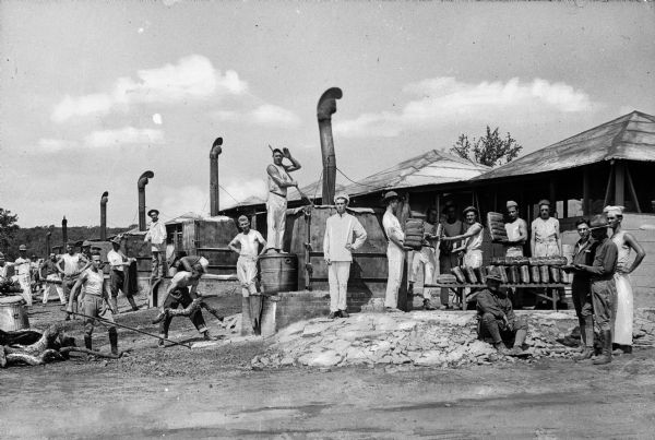 A view of men baking bread in a number of outdoor ovens at Camp Travis, named in 1917. On the right, men stand by a table with many loaves of bread, and men on the left work with the ovens.