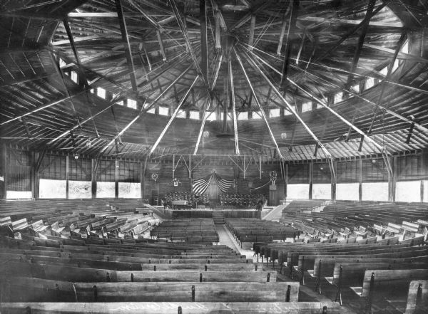 Interior of an auditorium. Wooden benches surround a platform where two United States flags are visible. On the ceiling, wooden beams make up the circular form of the building.
