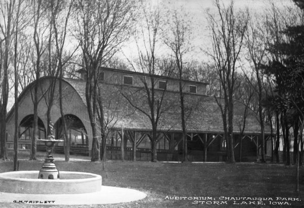 View of an open-sided auditorium and a fountain at Chautauqua Park, an establishment founded in 1874. The arched building stands in a wooded area. Caption reads: "Auditorium, Chautauque Park Storm Lake, Iowa."