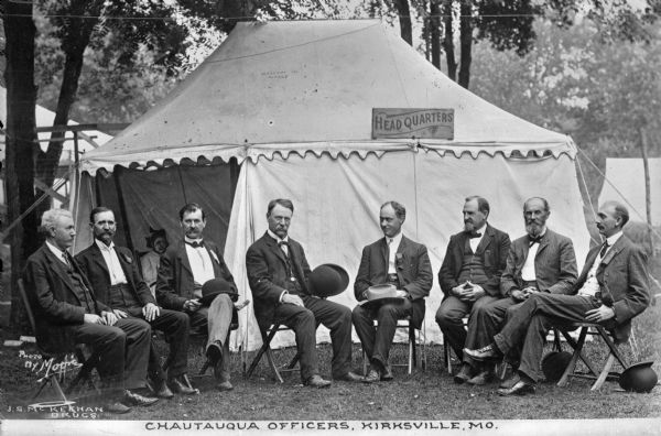View of eight men sitting outside a tent at Chautauqua Grounds, an establishment founded in 1874. A woman sits in the tent labeled, "Headquarters." Caption reads: "Chautauqua Officers, Kirksville, MO."