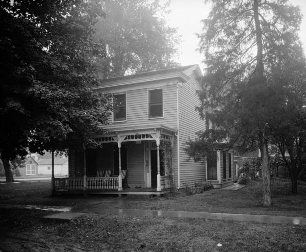 Exterior of Psi Beta Epsilon Fraternity House. Chairs are on the front porch. Trees surround the house, and a sidewalk leads to the door.