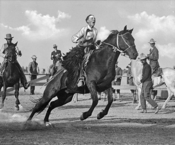 A man is holding his hat while riding a bucking bronco. Other men are following him on horseback at a "Frontier Days" rodeo. Observers look on from behind a wooden fence in the background.