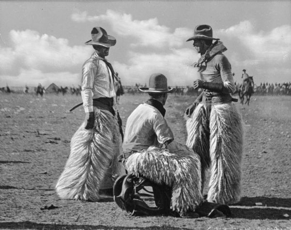Three cowboys wearing sheepskin chaps convene on the field at a "Frontier Days" rodeo. In the background are observers on horseback.