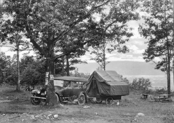 Sunset Camping Grounds View of an automobile and a pop-up camper set up near a lake at Sunset Camping Grounds. Labeled, "Chenango Trailer, Norwich, NY," the pop-up trailer company began manufacturing in 1920. Signs posted on a nearby tree encourage to "Keep in Line!" and advertise a dancing pavilion. Picnic benches and a fire pit can be seen on the campgrounds.