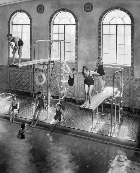 Elevated view of men and women preparing to jump from diving boards into an indoor pool at London Terrace Hotel. Other people are sitting on on the pool's edge. There are arched windows decorated with tile along the back wall.