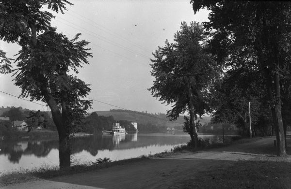 View of the Muskingum River. An excursion boat is moving past a dirt road in the foreground, and homes are in the distance.