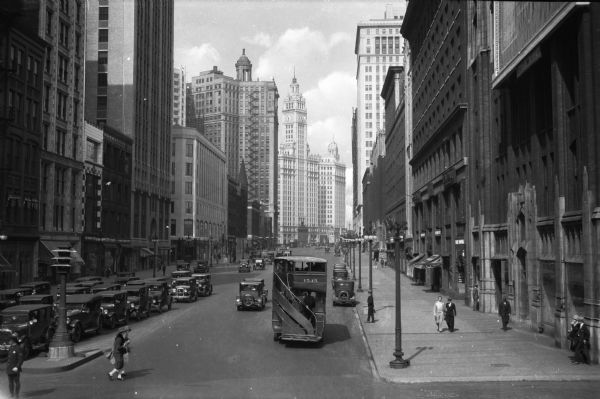 Slightly elevated view down Upper Michigan Boulevard. Pedestrians, automobiles, and a double-decker bus can be seen among tall buildings.