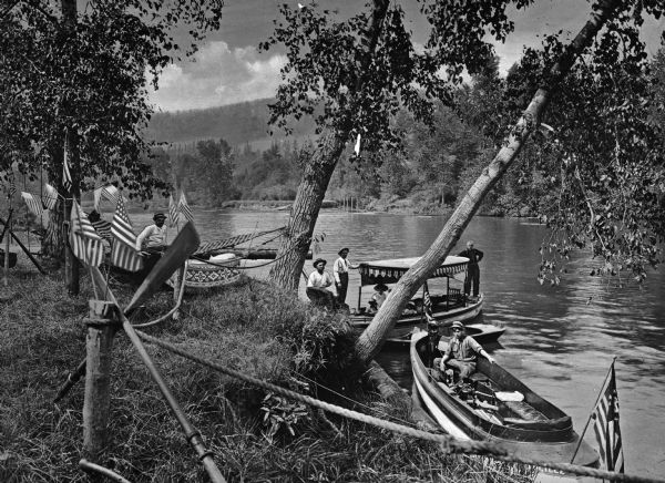 View from the wooded banks of a lake toward excursion boats in the water at the shoreline. Men pose near the boats and American flags decorate the foreground.
