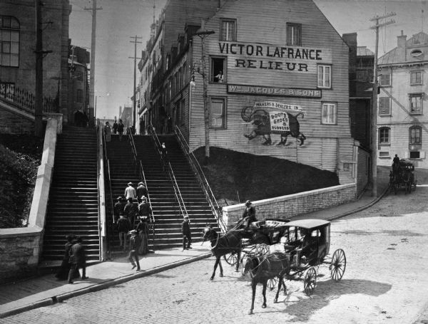 Men and women ascend a staircase toward a shoe store as horses and carriages travel along the street below. The sign on the store reads, "Victor LaFrance Relieur. Wm. Jacques & Sons. Makers & Dealers In Boots, Shoes, & Rubbers."