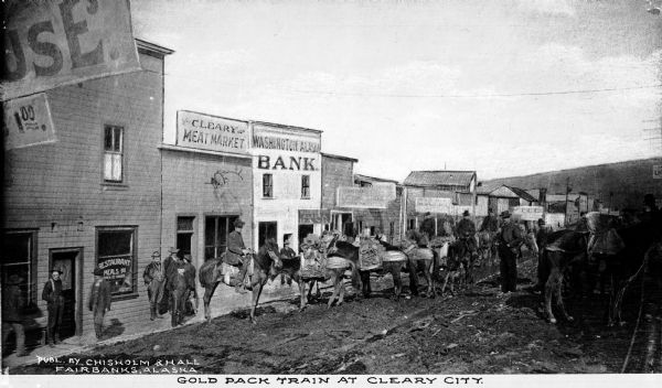 View of a gold pack train outside Washington Alaska Bank and Bleary Meat Market.  Published by Chisholm & Hall, Fairbanks, Alaska.