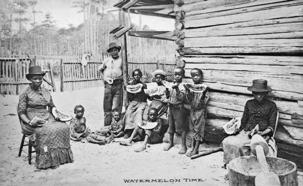 View of a family posed with slices of watermelon sitting in the yard near a log cabin. There is a large mortar and pestle in the right foreground.